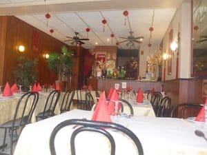 Salle de restaurant décorée dans un style asiatique, avec nappes claires, serviettes rouges pliées, statues traditionnelles et lanternes suspendues.