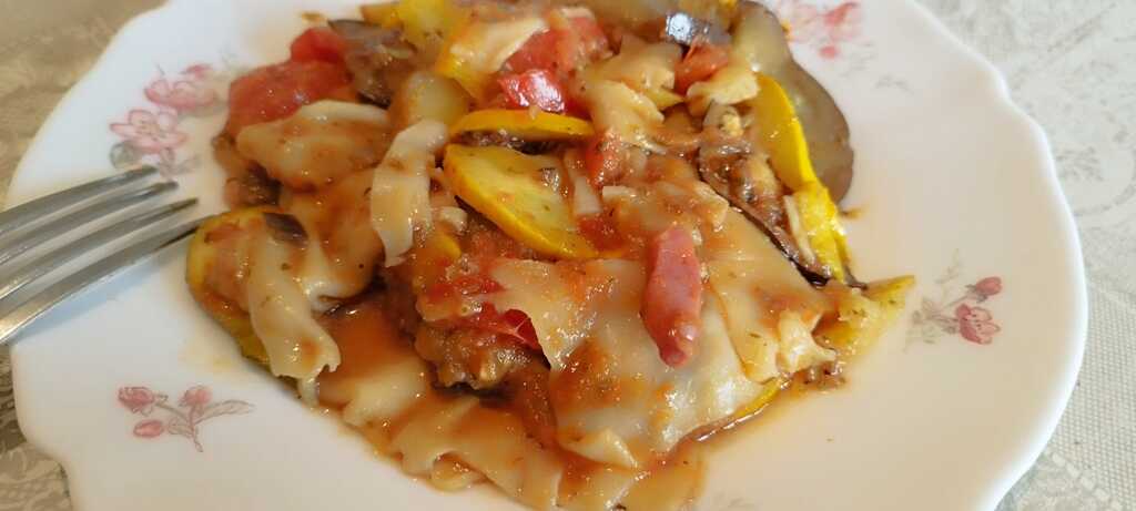 Gratin de courgettes jaunes, aubergines et tomates, nappé de fromage fondu, servi sur une assiette à motif floral avec une fourchette sur le côté.